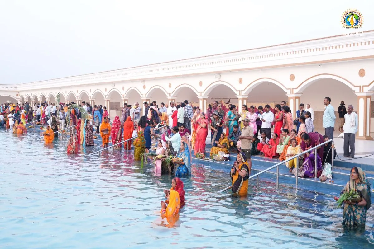 Devotees in the water of Divya Sarovar at Nurmahal Ashram during Chhath Puja 2025, a white arched colonnade beyond