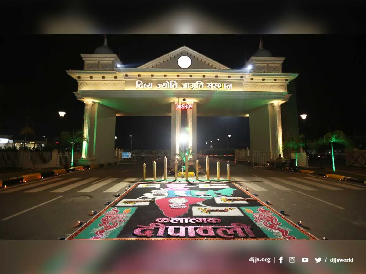 The entrance gate of Nurmahal Ashram lit at night for Diwali, with a large rangoli and earthen lamps on the road before it